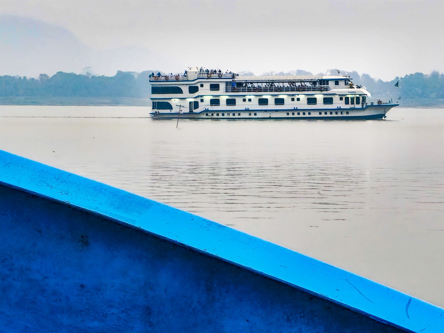 Multi-deck river cruise ship sailing a broad, misty river in northeast India