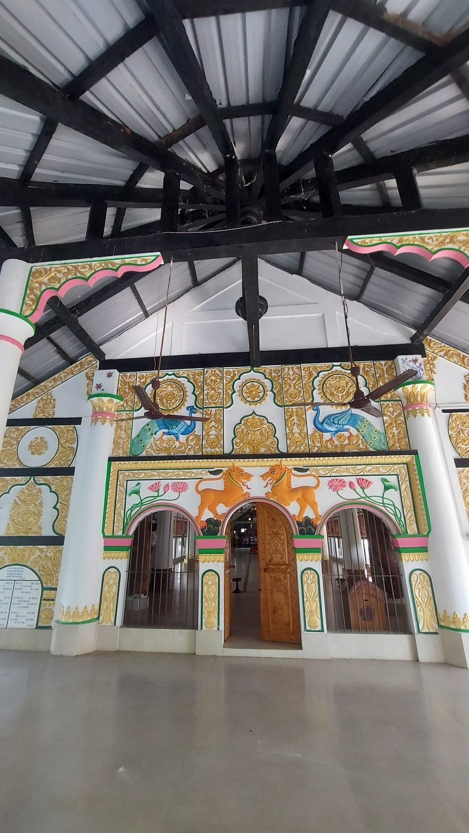 Ornate white and gold facade with painted peacocks and lions inside an open prayer hall in Assam